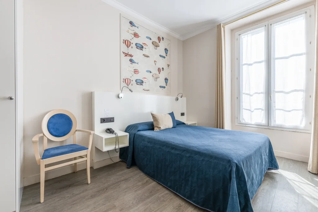 Chambre double avec lit au couvre-lit bleu, fauteuil médaillon et grande fenêtre voilée