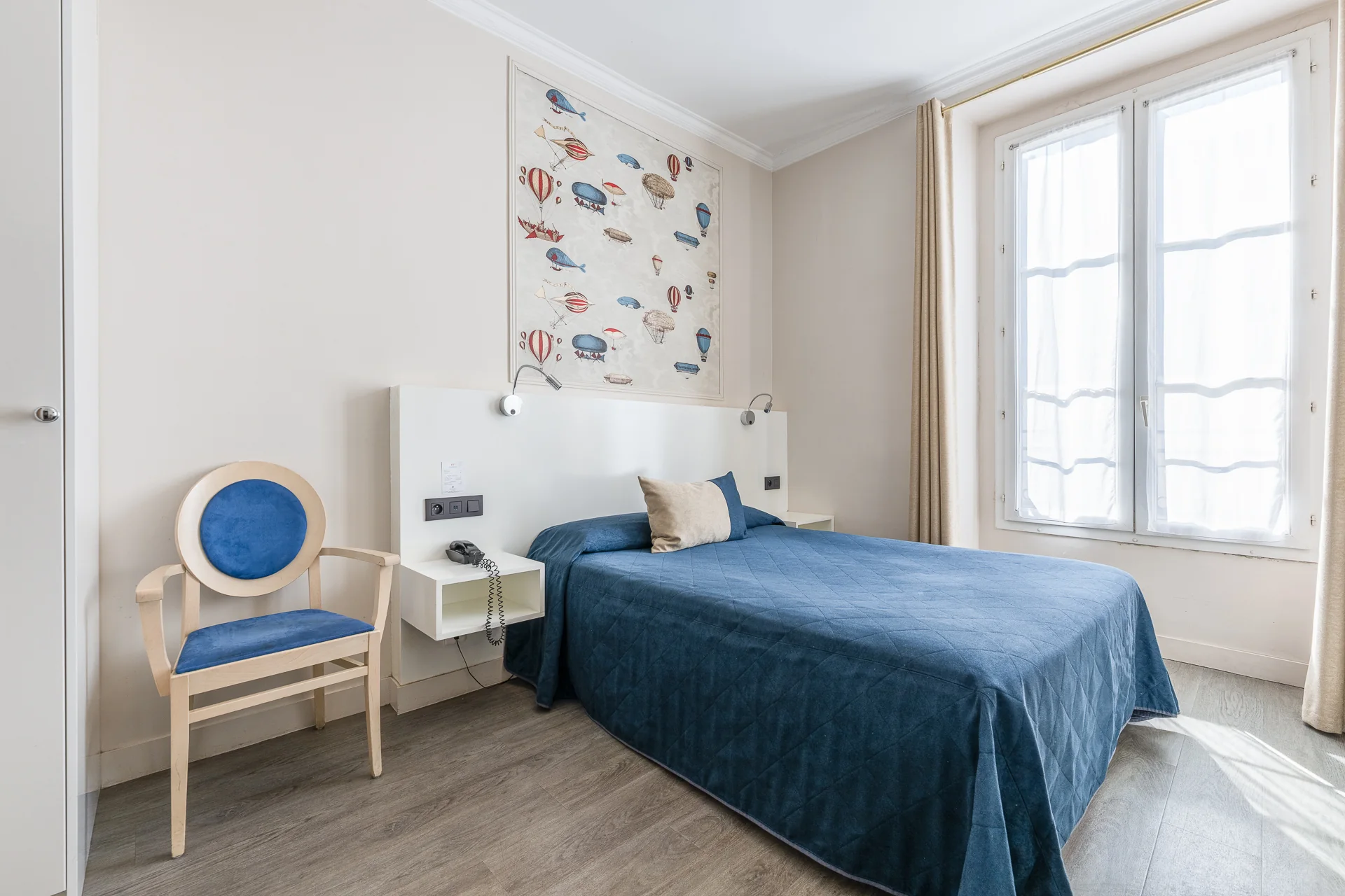 Chambre double avec lit au couvre-lit bleu, fauteuil médaillon et grande fenêtre voilée