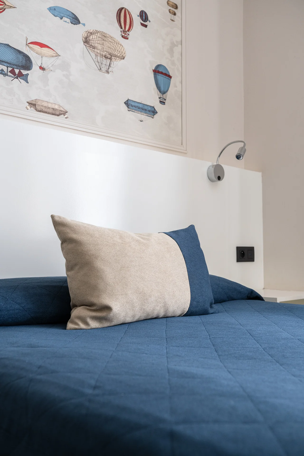 Coussin beige et bleu sur un lit, sous une liseuse et un panneau de montgolfières