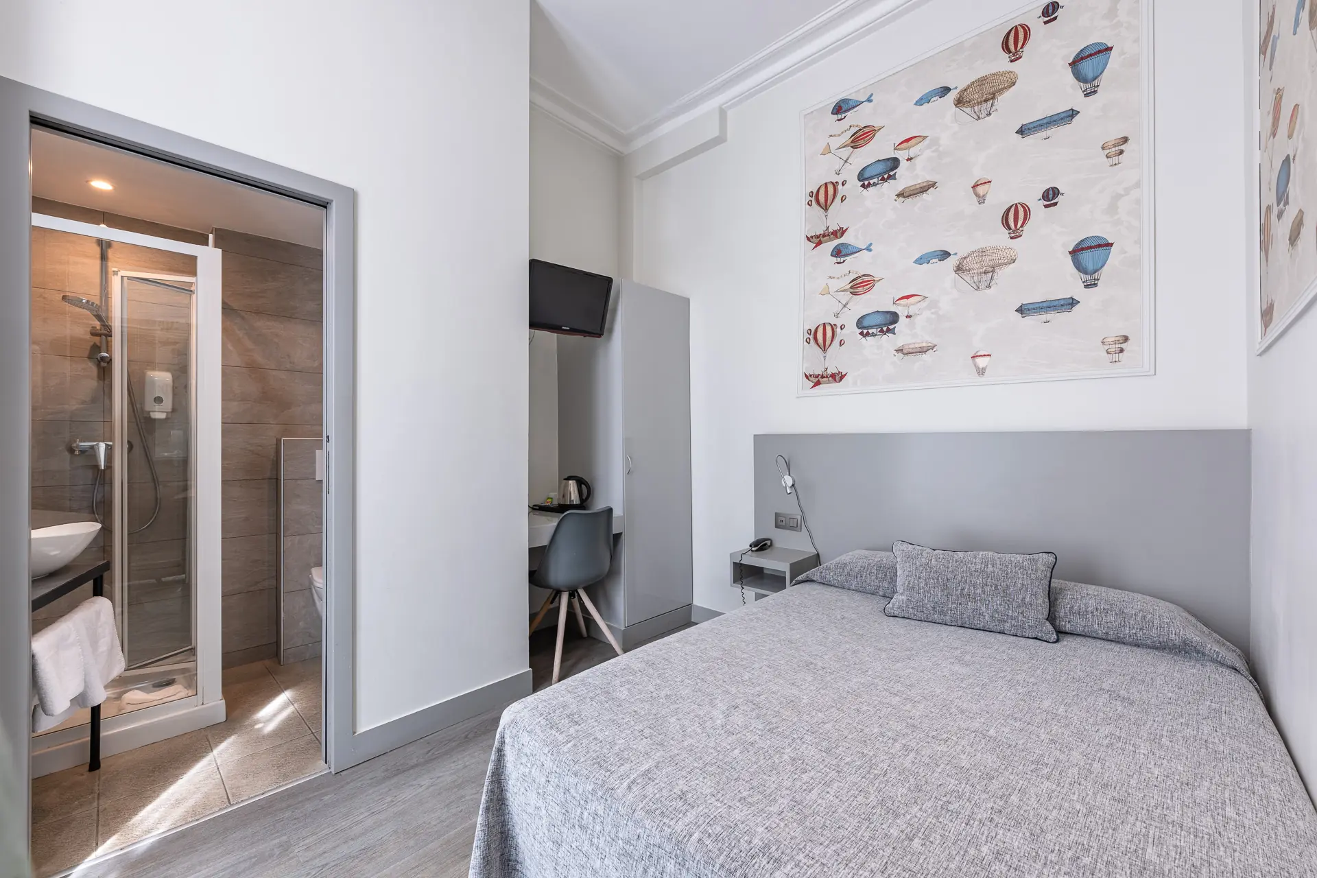 Chambre single avec couvre-lit gris, tableau de montgolfières et salle de douche ouverte