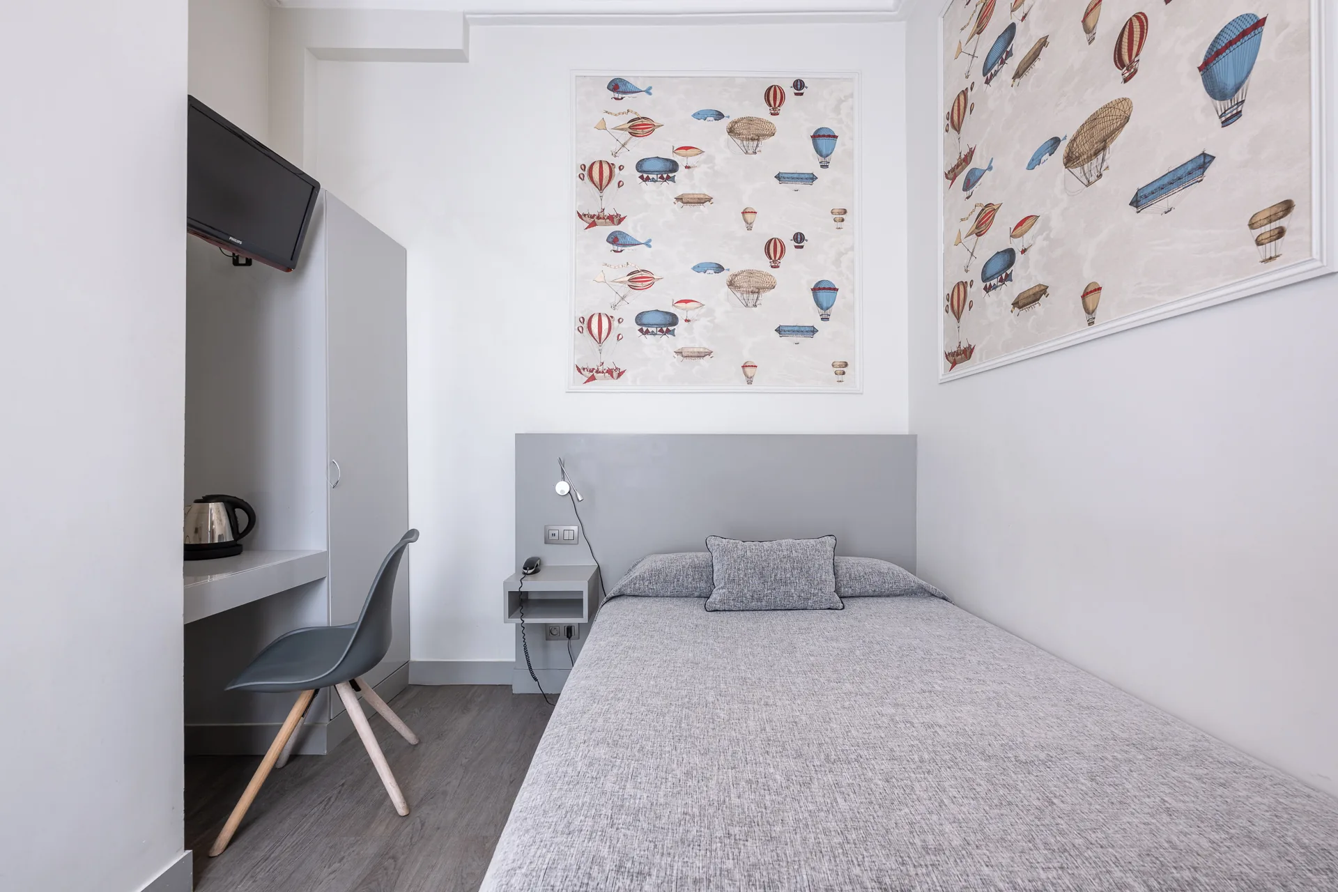 Chambre single avec lit au couvre-lit gris, coin bureau, bouilloire et télévision murale