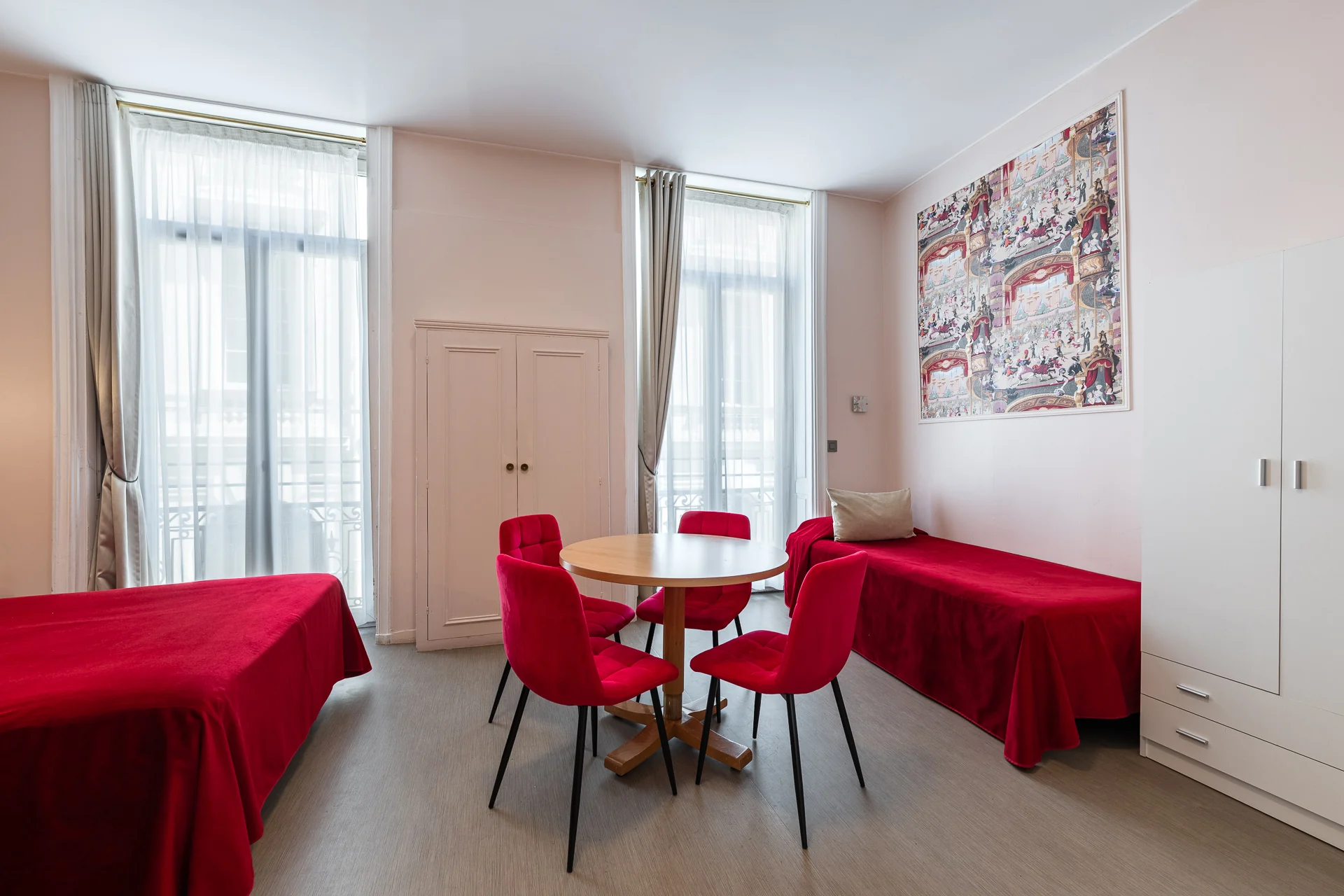 Table ronde et chaises rouges entre deux lits, devant des portes-fenêtres voilées