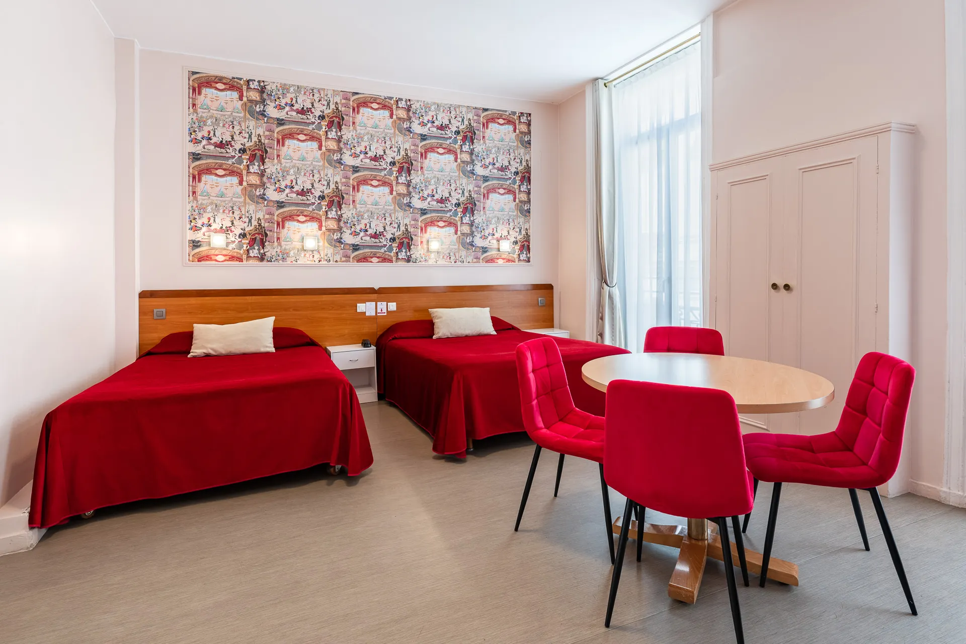 Chambre triple avec deux lits rouges, table ronde, chaises rouges et armoire blanche