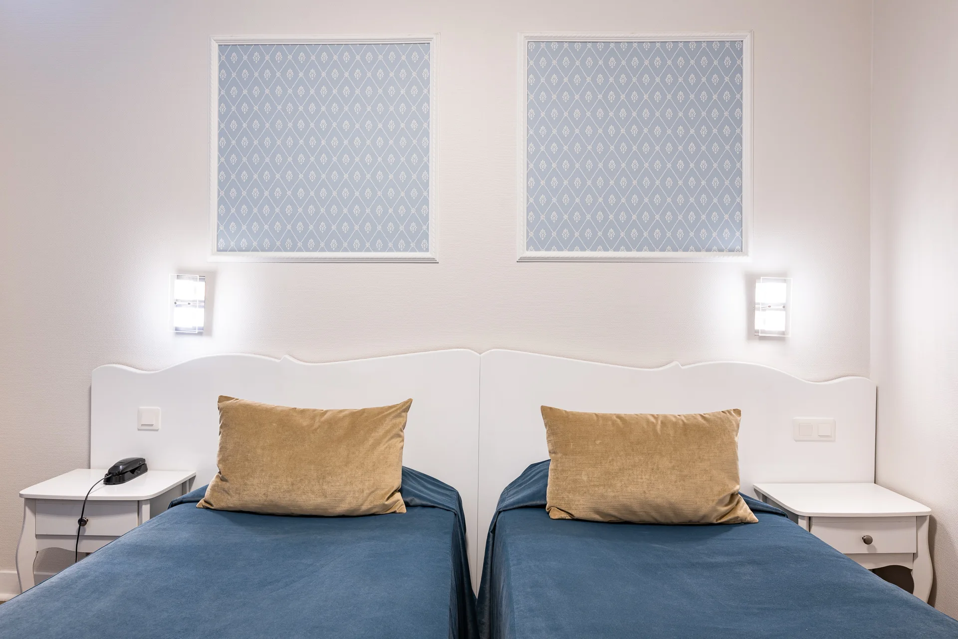 Deux lits simples aux couvre-lits bleu canard et coussins beiges, sous deux appliques